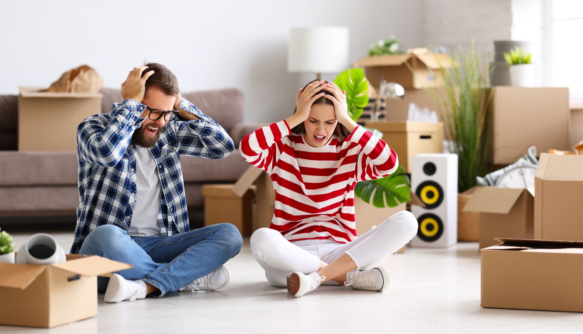 Young couple sitting on the floor of their new home, looking stressed with their hands on their heads, surrounded by moving boxes.