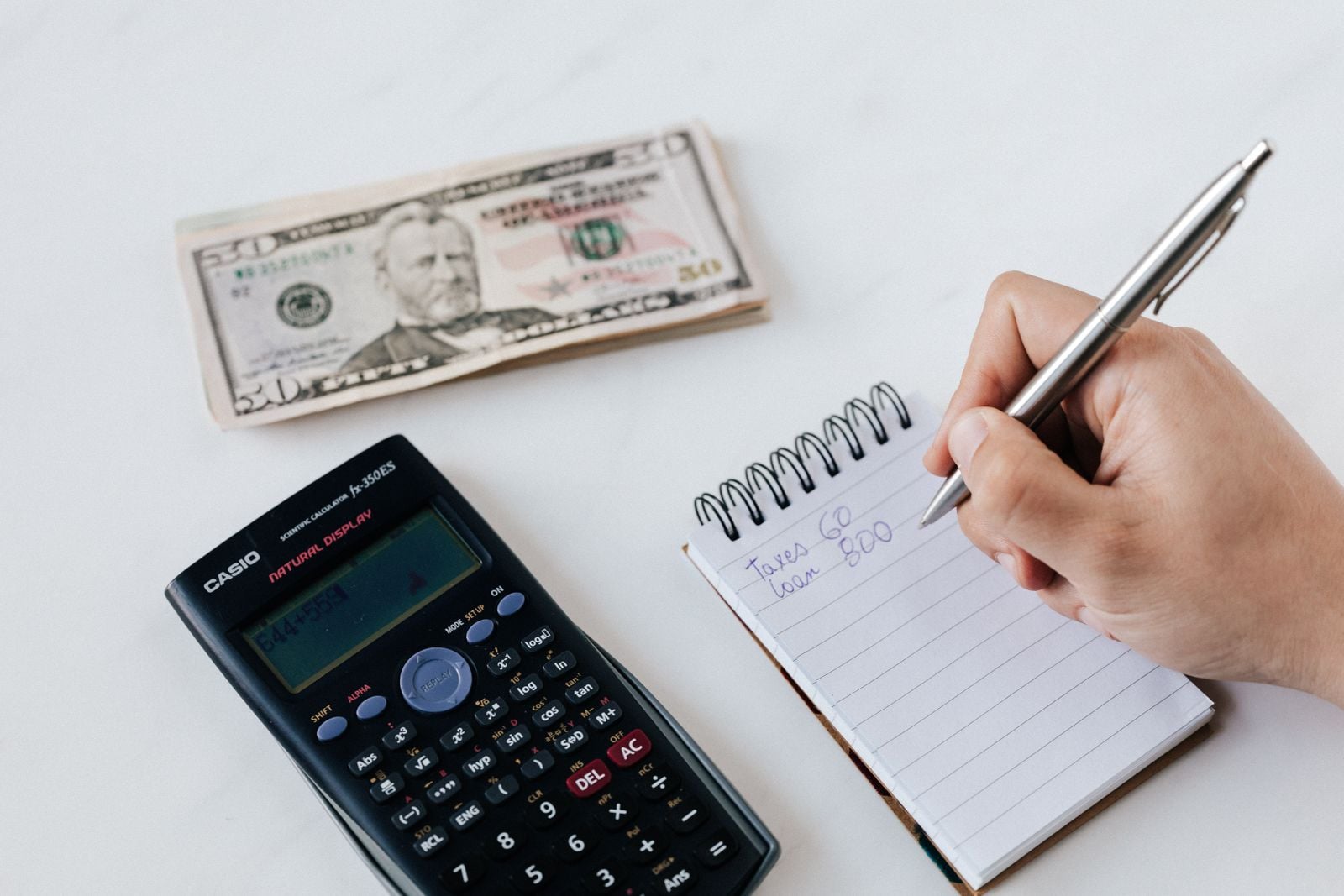 Budget review with calculator and notebook Person writing in a notebook beside a calculator and cash.